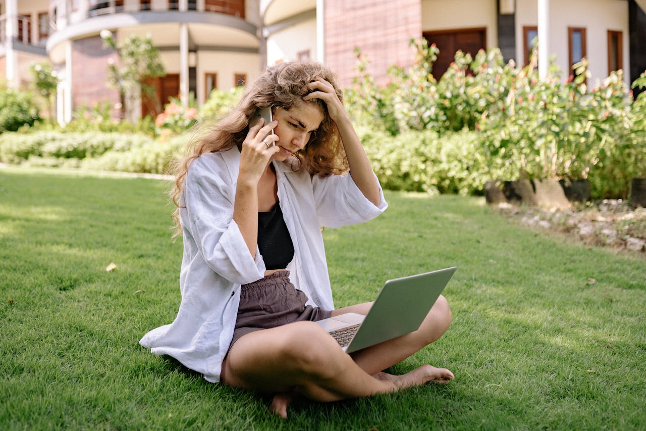 services-09 A woman sits on a lawn, using a laptop and phone, working remotely outdoors in a tropical setting.